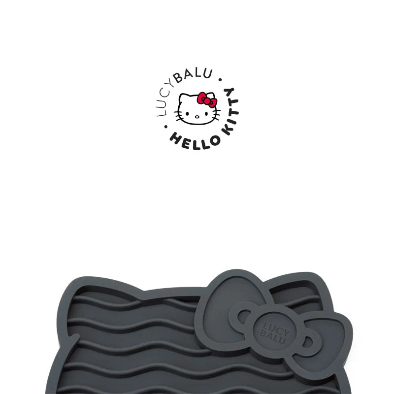 Graue Schleckmatte aus Silikon in Hello-Kitty-Kopfform mit Wellenrillen und Schleife mit LucyBalu-Prägung, darüber das runde Logo LucyBalu x Hello Kitty.
