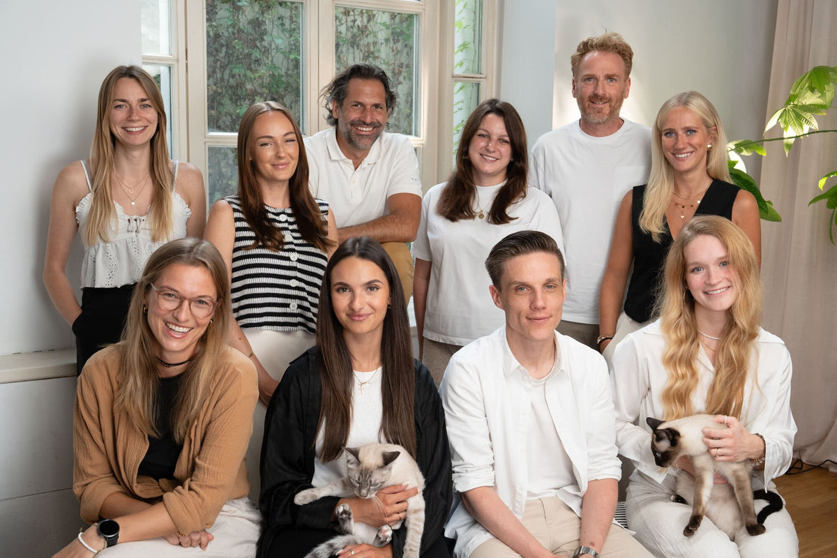 Das LucyBalu-Team 2025: zehn lächelnde Personen in heller Kleidung in einem Raum mit großen Fenstern, zwei davon halten Siamkatzen auf dem Arm.