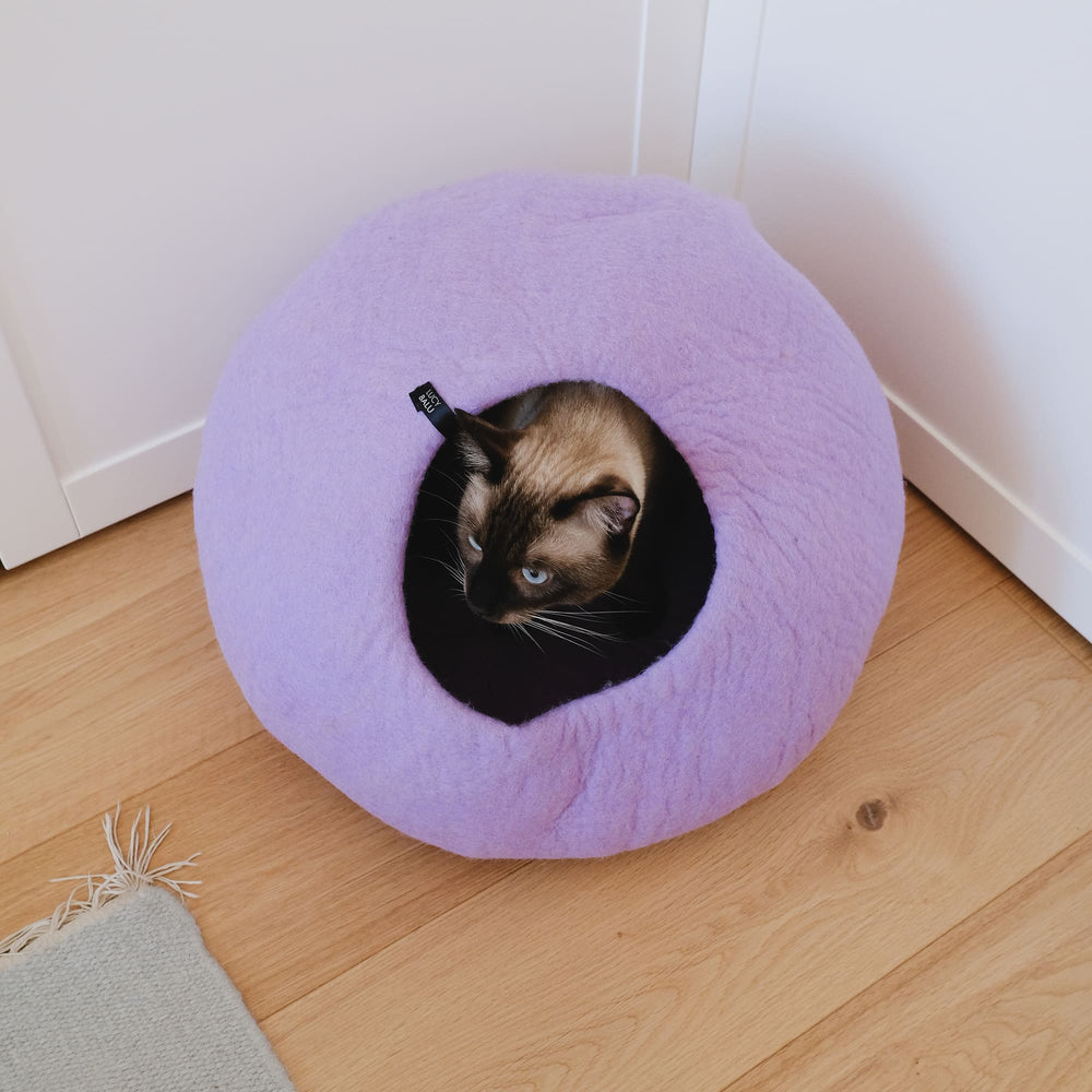 PEBBLE felt cat cave in lilac on a wooden floor seen from above, a Siamese cat looks out of the entrance
