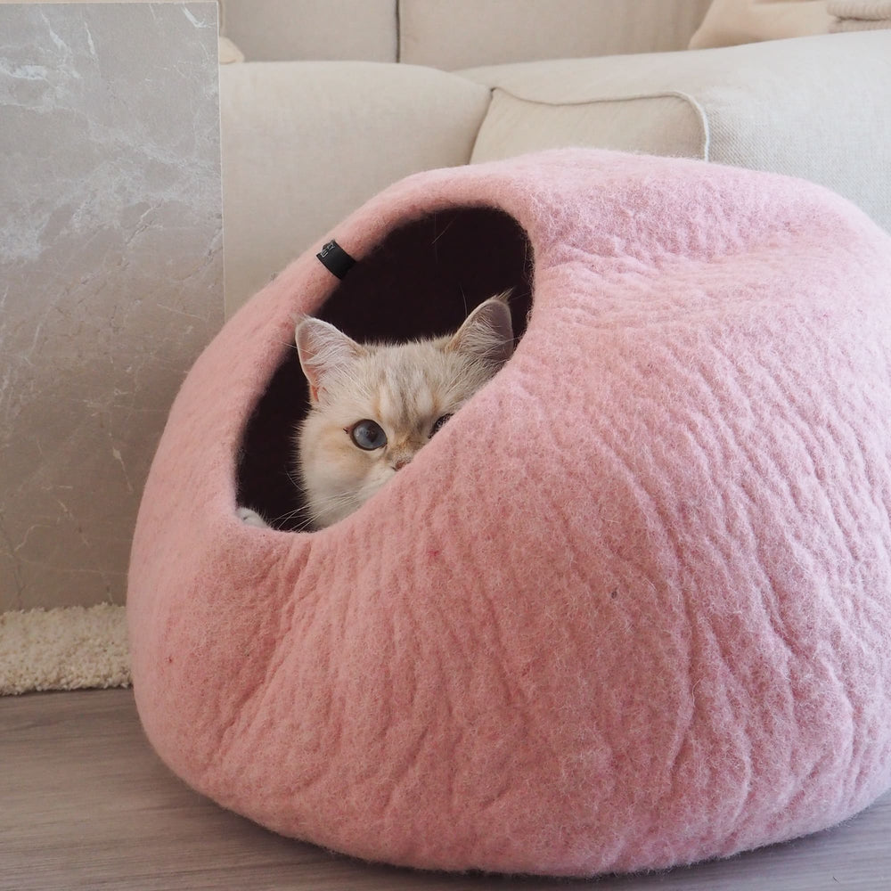 PEBBLE felt cat cave in pink with a light-coloured cat peeking out of the opening, in front of a sofa