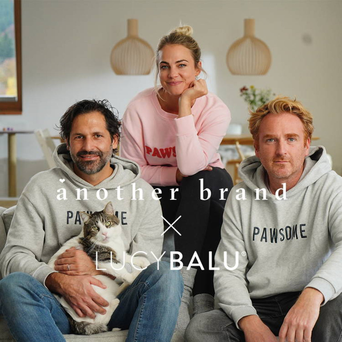 Zwei Männer in grauen „Pawsome“-Hoodies und eine Frau in rosa Sweatshirt sitzen auf einem Sofa, einer hält eine getigerte Katze mit weißem Bauch, darüber das Logo another brand x LucyBalu.
