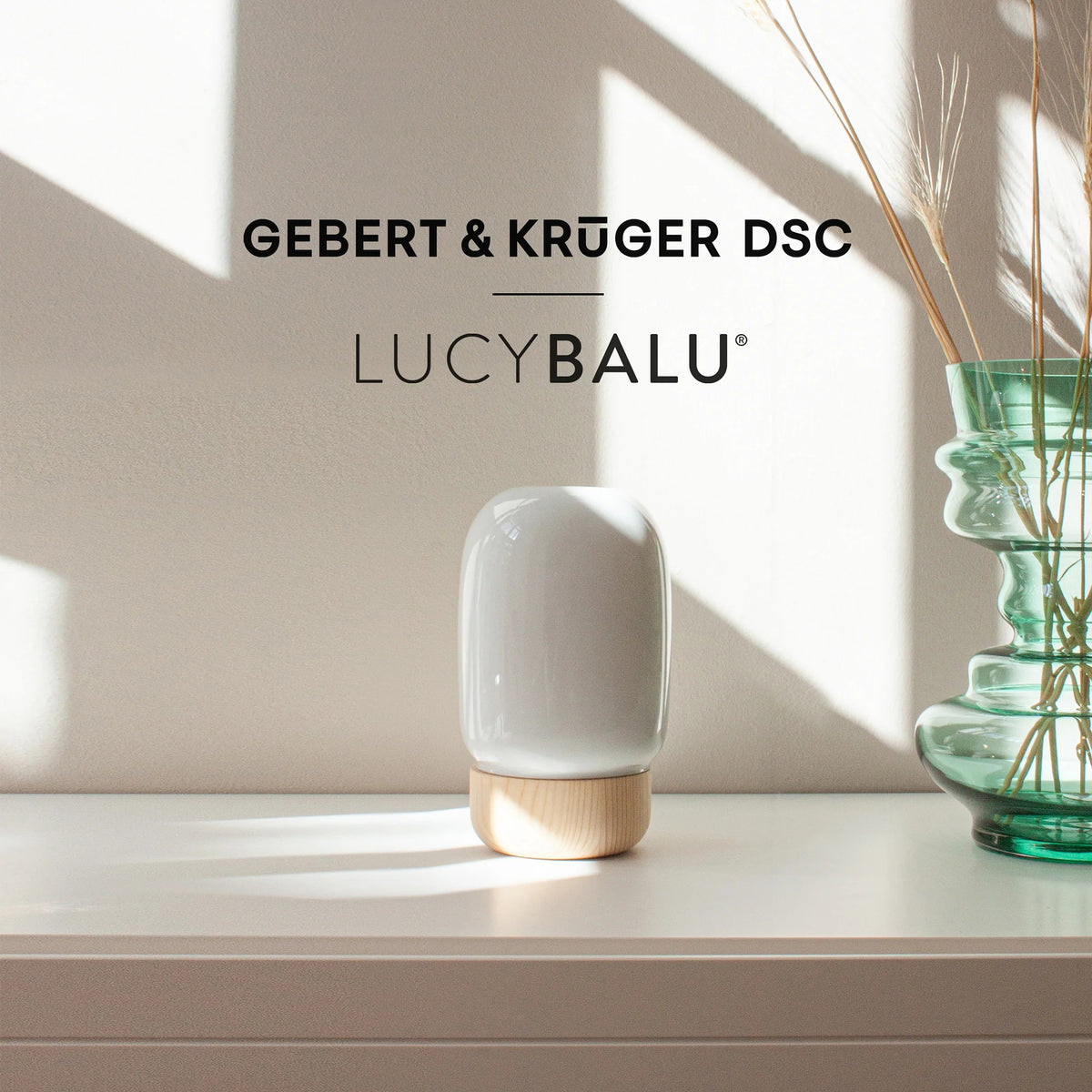 Weiße Katzenurne mit Holzsockel auf einem hellen Sideboard im Sonnenlicht, daneben eine grüne Glasvase mit Trockengräsern und darüber das Logo Gebert & Krüger DSC x LucyBalu.