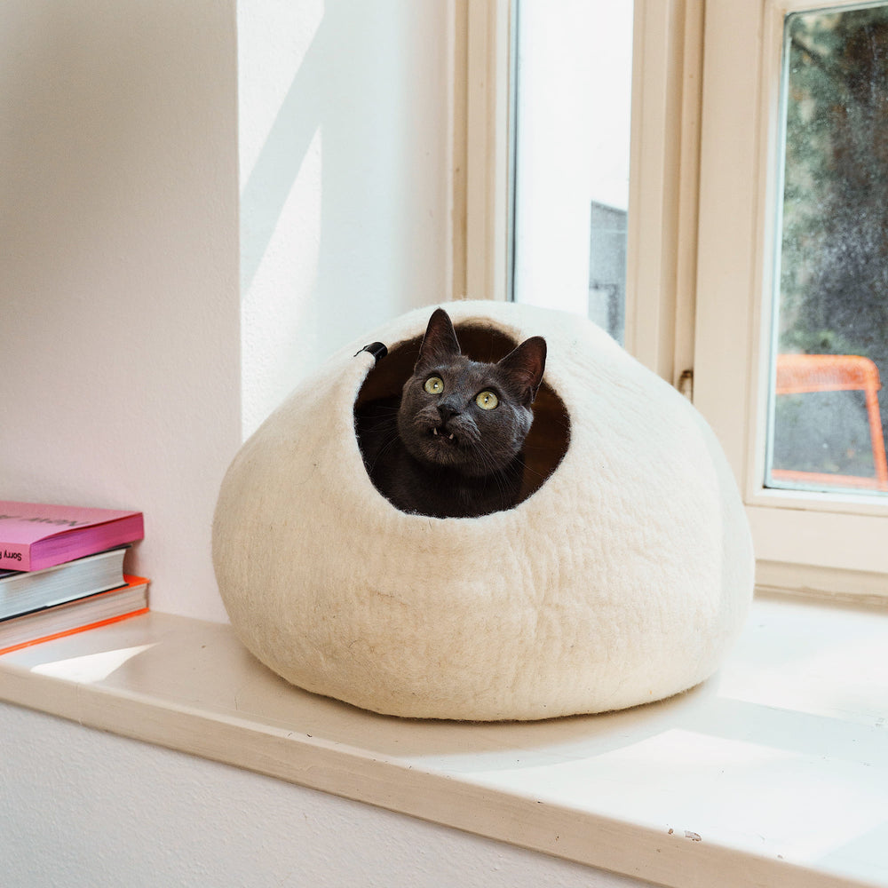 PEBBLE felt cat cave in cream white on a windowsill, a grey cat looks up from the entrance
